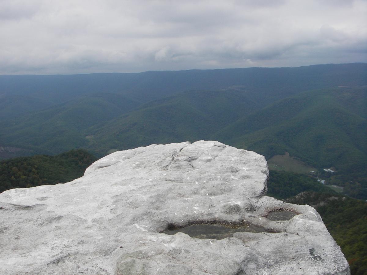 A rocky outcrop overlooking a vast green landscape of rolling mountains under an overcast sky. The foreground features a large, flat stone surface, while the background showcases layers of hills fading into the distance. The scene conveys a sense of nature's beauty and tranquility. North Fork Mountain Trail mountain bike trail.