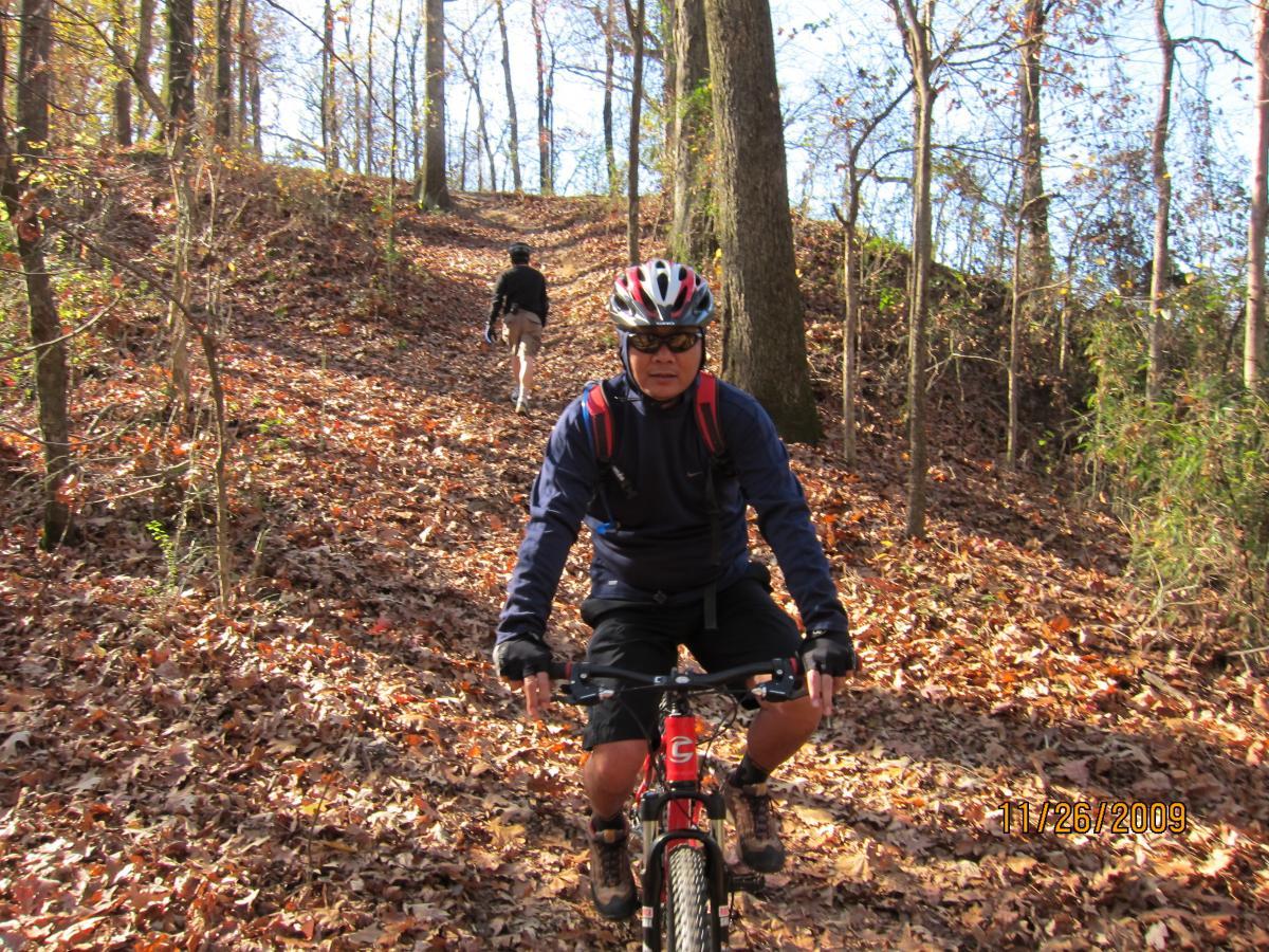 A person riding a mountain bike on a wooded trail covered in autumn leaves, with another individual walking uphill in the background. The scene is bathed in natural sunlight, highlighting the vibrant colors of the foliage and the tranquil atmosphere of the outdoor setting. Stanky Creek mountain bike trail.