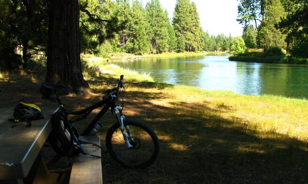 A black mountain bike rests against a wooden bench near a tranquil river, surrounded by lush green trees and grass under bright sunlight. The peaceful setting invites outdoor activities and relaxation. Deschutes River mountain bike trail.
