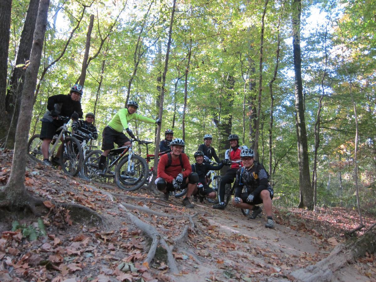 A group of eight mountain bikers poses for a photo on a forest trail, surrounded by trees with green leaves and fallen autumn foliage. They are wearing helmets and biking gear, with some bikes parked nearby. The scene conveys a sense of camaraderie and outdoor adventure. Stanky Creek mountain bike trail.