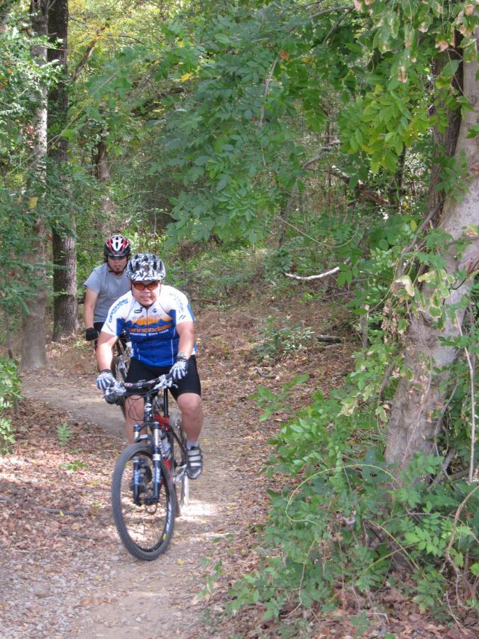 Two mountain bikers riding on a gravel trail surrounded by trees and foliage. One cyclist, wearing a blue jersey and a helmet, is in the foreground, while another cyclist, dressed in gray, follows behind. The scene captures a peaceful outdoor setting with autumn leaves. Stanky Creek mountain bike trail.