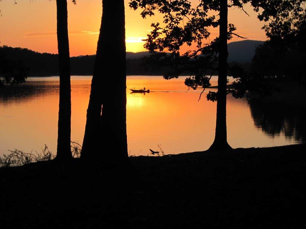 A serene sunset over a calm lake, with silhouettes of trees framing the scene and a small boat gliding across the water. The sky is filled with warm hues of orange and yellow, reflecting on the tranquil surface of the lake. Lovit: Section 1 mountain bike trail.