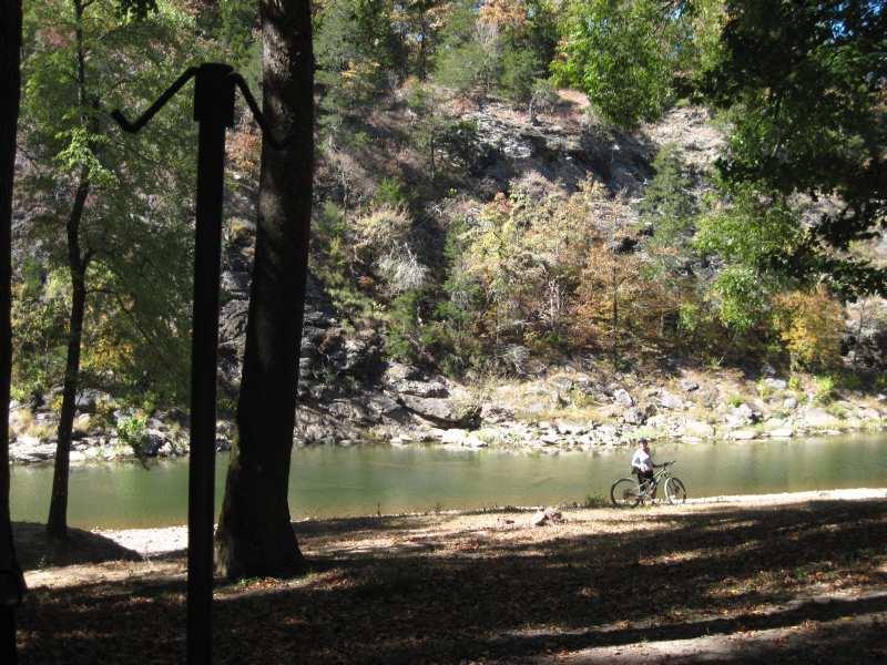 A tranquil riverside scene featuring a cyclist resting beside a calm river, surrounded by lush trees and a rocky hillside. The area displays autumn foliage, creating a picturesque outdoor atmosphere. Womble mountain bike trail.