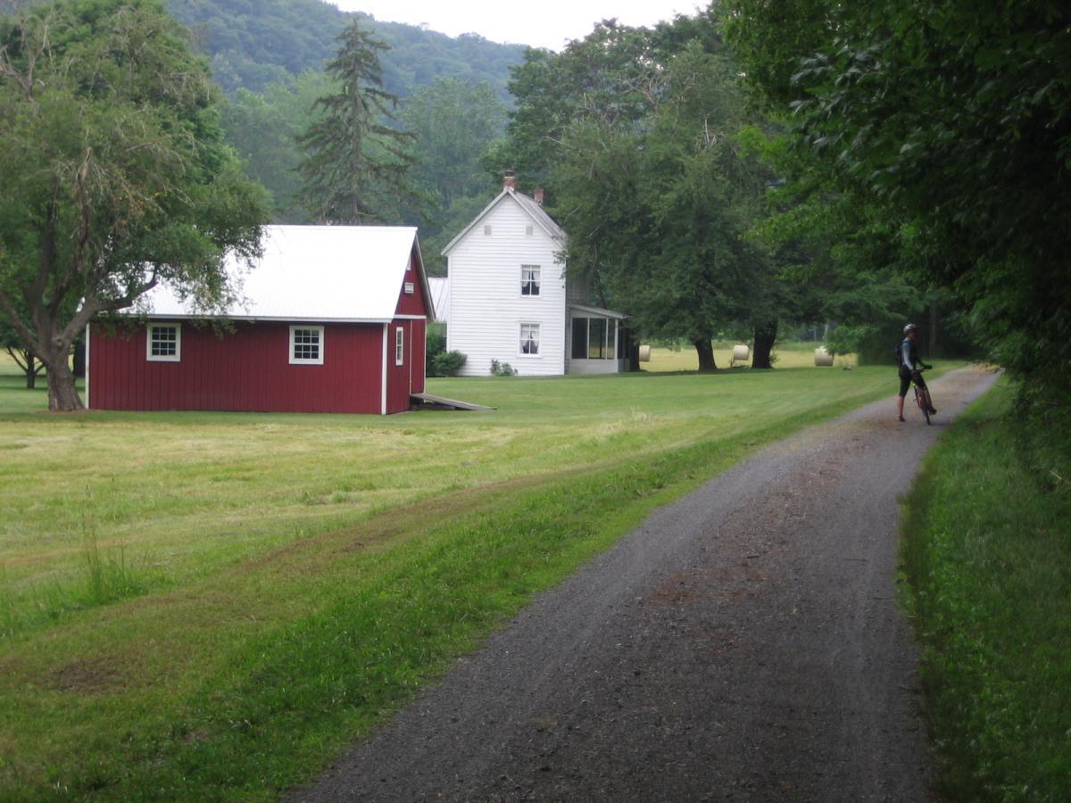 A scenic view of a rural landscape featuring a red barn and a white house surrounded by lush green grass and trees. A gravel pathway runs alongside the property, where a person is seen standing with a bicycle. In the background, hay bales can be spotted in the field. Greenbrier River Trail mountain bike trail.