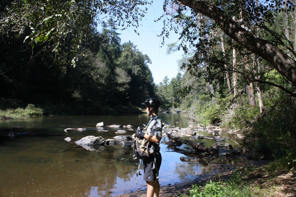 A person in a cycling helmet and gloves stands beside a calm river, gazing at the water. The scene is surrounded by lush greenery and trees, with sunlight filtering through the branches. Rocks partially submerged in the river are visible, creating a serene outdoor atmosphere. River Loop mountain bike trail.