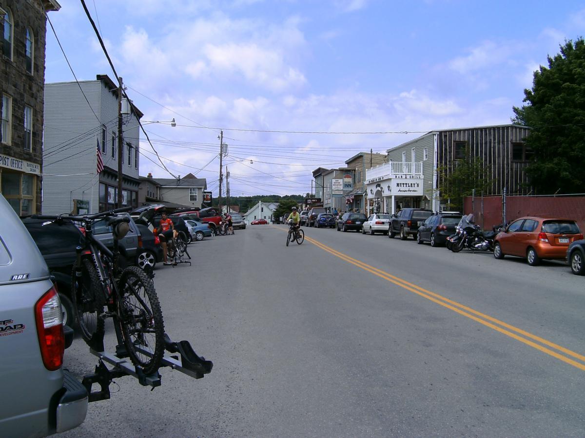 A street view of a small town, featuring parked cars on both sides of a wide road. Bicycles are mounted on the back of a vehicle in the foreground. People are gathered near the vehicles, with one cyclist riding down the road. The buildings lining the street include shops and a post office, under a partly cloudy sky. CVI Trails mountain bike trail.