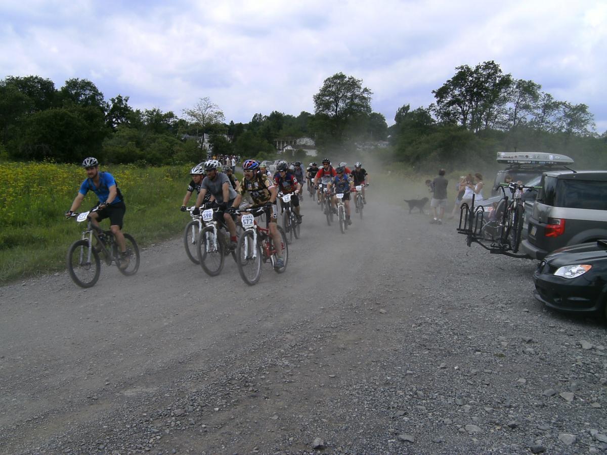 A group of cyclists in helmets and biking attire participate in a mountain bike race on a gravel path, creating a cloud of dust behind them. In the background, spectators watch from the sidelines, while some vehicles are parked nearby. Lush greenery and trees line the sides of the path under a partly cloudy sky. CVI Trails mountain bike trail.