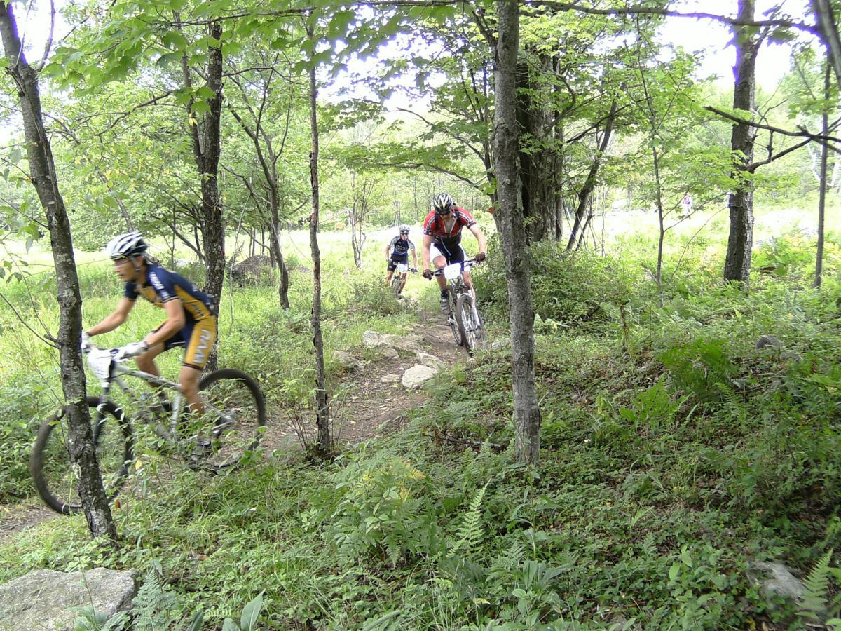 Mountain bikers navigating a narrow, rocky trail surrounded by lush greenery and trees. The scene captures three cyclists in motion, showcasing the excitement and challenge of off-road biking in a natural environment. CVI Trails mountain bike trail.