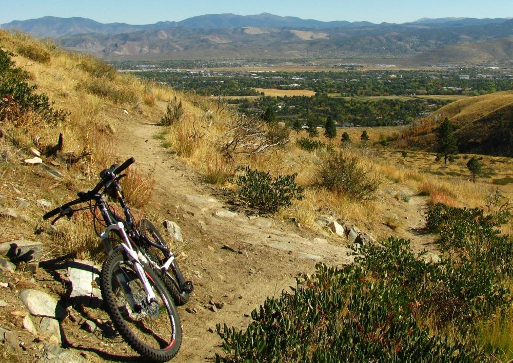 A mountain bike resting on a dirt trail overlooking a valley with green trees and fields, framed by distant mountains under a clear blue sky. Ash Canyon Trails mountain bike trail.