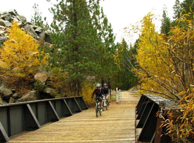 Two cyclists riding on a wooden bridge surrounded by autumn foliage, with vibrant yellow leaves and evergreen trees in a scenic outdoor setting. Bizz Johnson Trail mountain bike trail.
