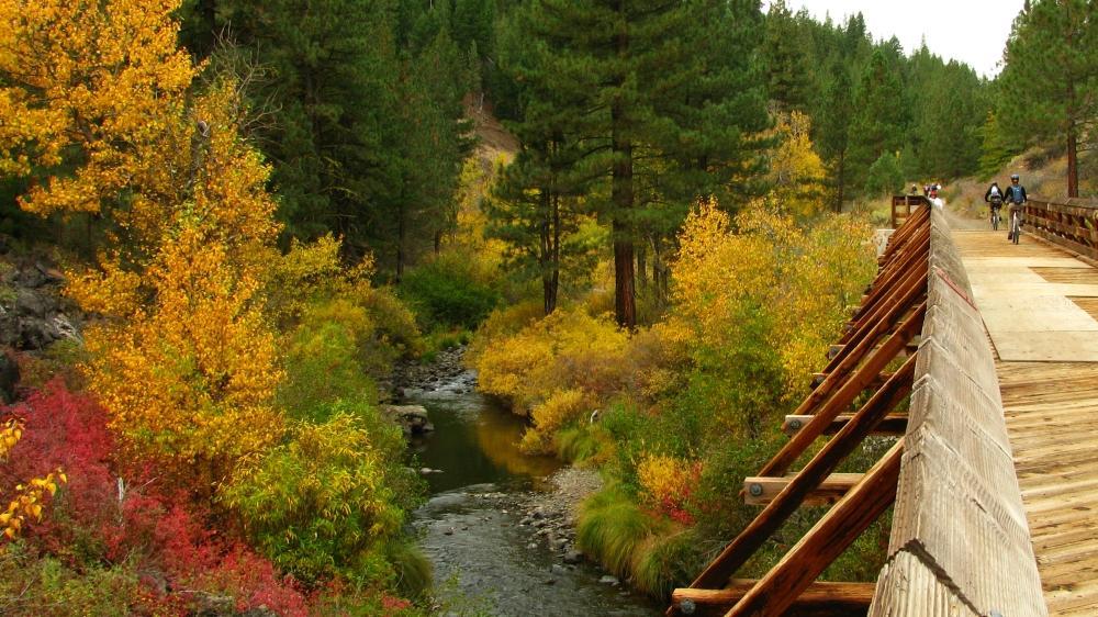 A scenic view of a wooden bridge over a winding creek, surrounded by vibrant autumn foliage in shades of orange, yellow, and red. Tall pine trees and rolling hills are visible in the background, creating a picturesque natural landscape. Two cyclists traverse the bridge, adding a sense of activity to the serene environment. Bizz Johnson Trail mountain bike trail.