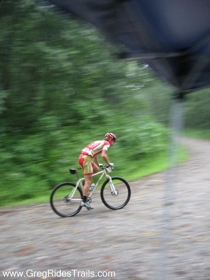 A cyclist wearing a red and white jersey rides rapidly on a gravel trail, surrounded by lush green foliage. The image conveys a sense of motion and speed, capturing the dynamic nature of mountain biking in a natural setting. Winding Stairs Loop mountain bike trail.