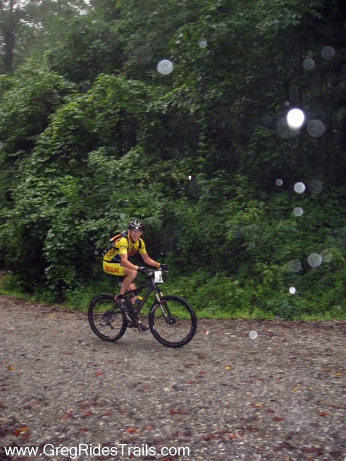 A mountain biker in a yellow jersey rides along a gravel trail in rainy weather, surrounded by dense green foliage. Rain droplets are visible in the air as they pedal forward. Winding Stairs Loop mountain bike trail.