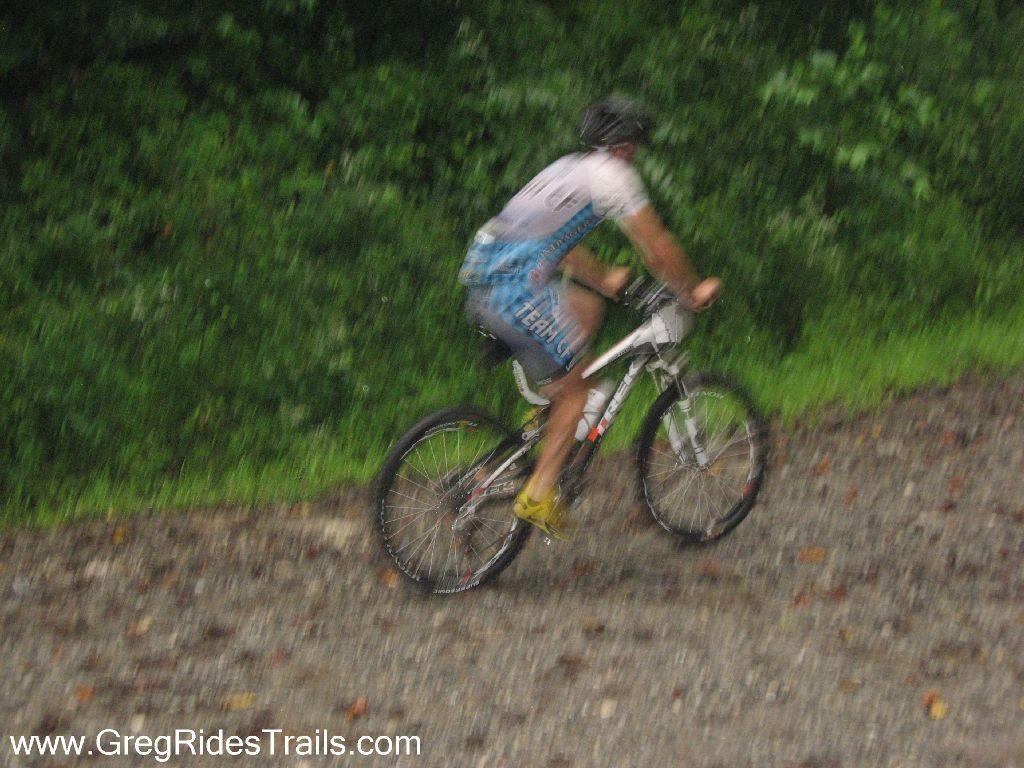 A blurred image of a person riding a mountain bike on a gravel path surrounded by greenery, suggesting movement through a natural environment. The cyclist is wearing a blue and white jersey and a helmet. The scene appears slightly rainy or misty, adding to the sense of action. Winding Stairs Loop mountain bike trail.