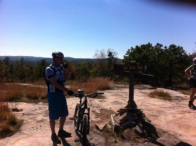 A mountain biker in a helmet and athletic attire stands next to a bicycle, holding a water bottle and looking at a trail sign that reads "Big Rock Trail." The surrounding landscape features rocky terrain and sparse vegetation under a clear blue sky, with rolling hills in the background. Another cyclist can be seen in the distance, also on the trail. DuPont State Recreational Forest mountain bike trail.
