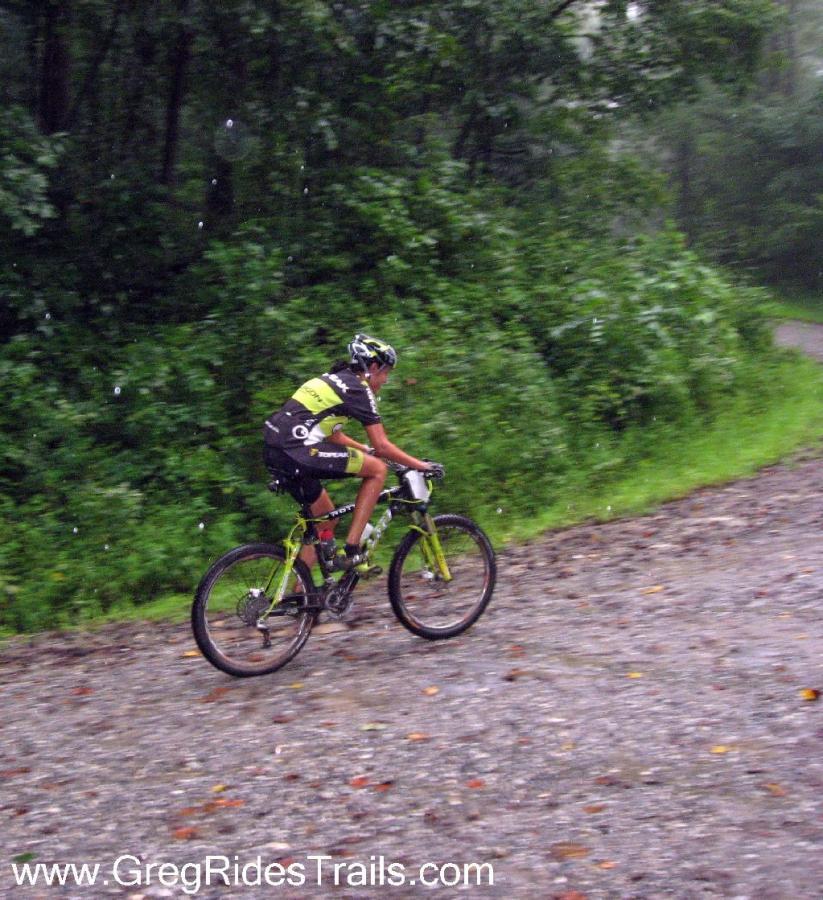 A cyclist riding a mountain bike along a gravel trail in a forested area, with raindrops visible in the air and lush greenery in the background. The cyclist is wearing a black and green racing outfit and a helmet. Winding Stairs Loop mountain bike trail.