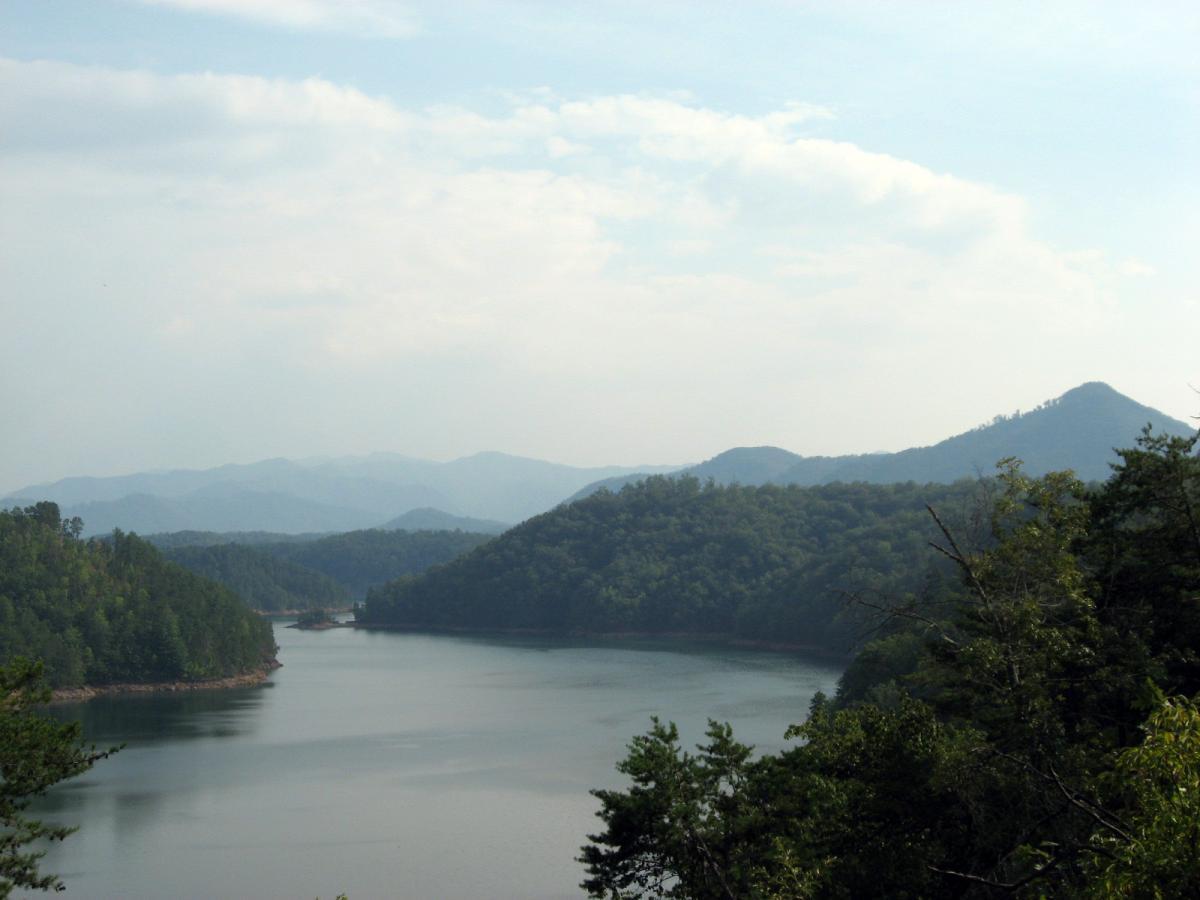 Scenic view of a calm lake surrounded by lush green hills and distant mountains under a cloudy sky. The water reflects the landscape, creating a tranquil and picturesque atmosphere. Tsali Recreation Area mountain bike trail.