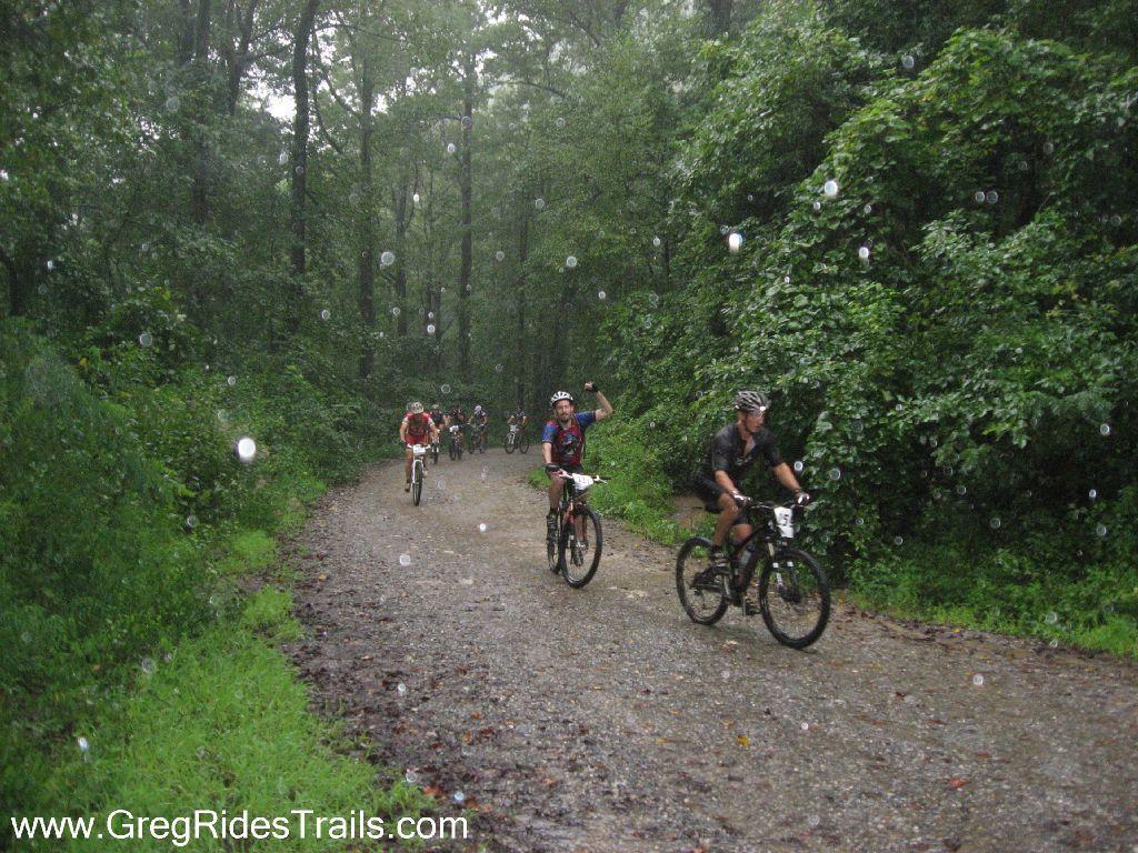 Mountain bikers riding on a gravel trail in a wooded area during rainfall, surrounded by lush greenery and tree cover. Some cyclists are smiling and waving at the camera, while others continue pedaling through the rain. Winding Stairs Loop mountain bike trail.