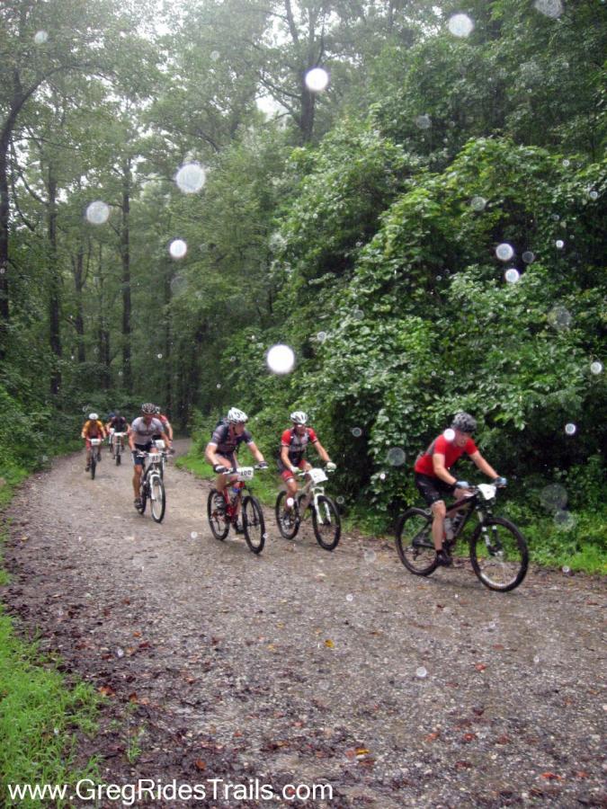 A group of mountain bikers racing along a gravel trail in a dense forest during rainy weather. The scene shows a lush green backdrop with trees, and raindrops are visible in the foreground, adding to the atmosphere of a wet and challenging ride. The cyclists are wearing helmets and racing gear, indicating a competitive event. Winding Stairs Loop mountain bike trail.