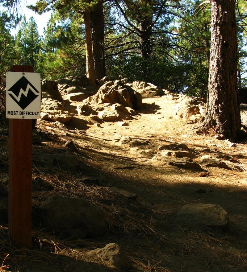 A rocky hiking trail labeled "Most Difficult," surrounded by pine trees and natural greenery, with sunlight illuminating the path. A signpost indicates the level of difficulty for hikers. Grand Slam mountain bike trail.