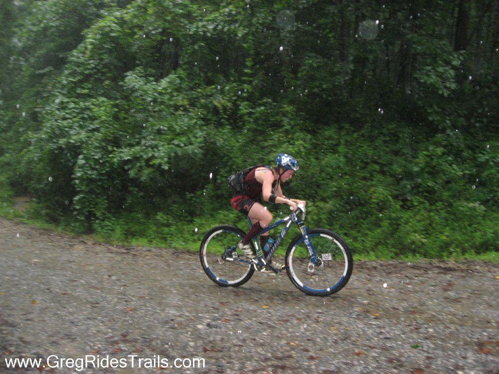 A person riding a mountain bike down a gravel path during a rainstorm, with greenery in the background. Water droplets are visible in the air, capturing the essence of a wet and adventurous ride. The cyclist is wearing a helmet and appears focused on navigating the terrain. Winding Stairs Loop mountain bike trail.