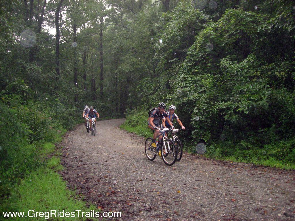 Alt text: Mountain bikers riding on a gravel path surrounded by dense greenery in a rain-soaked forest. Winding Stairs Loop mountain bike trail.