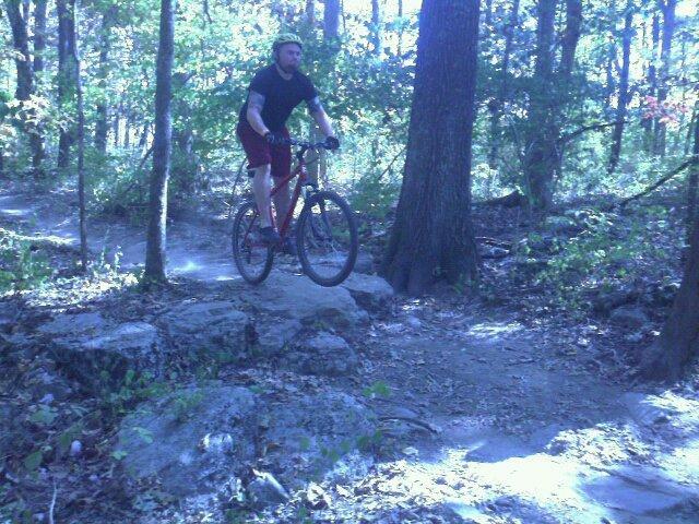 A mountain biker riding over a rocky trail in a wooded area, surrounded by tall trees and sunlight filtering through the leaves. The rider is wearing a black shirt and helmet, navigating the uneven terrain. Lock 4 mountain bike trail.