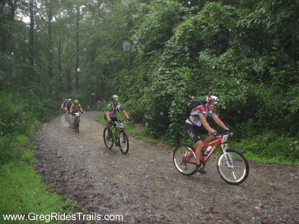 A group of mountain bikers riding on a gravel path through a dense, rain-soaked forest. The scene captures several riders in various colored jerseys and helmets, with raindrops visible in the air. Lush green foliage surrounds the trail, emphasizing the outdoor, adventurous atmosphere despite the rainy conditions. Winding Stairs Loop mountain bike trail.