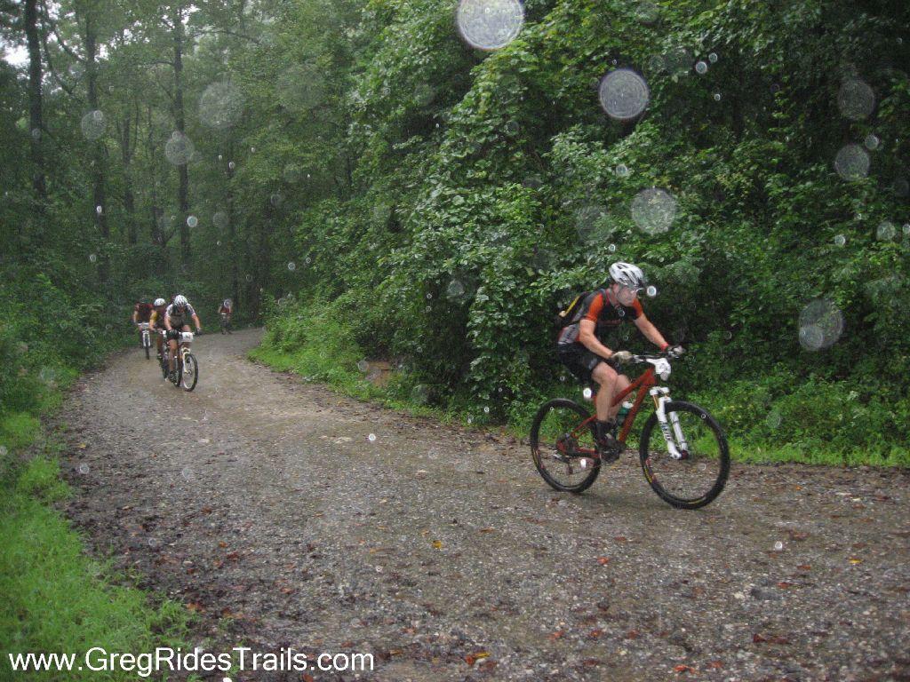 Mountain bikers riding on a gravel trail through a forest on a rainy day, with droplets visible in the air. Lush green trees surround the path, creating a natural setting filled with moisture. Winding Stairs Loop mountain bike trail.