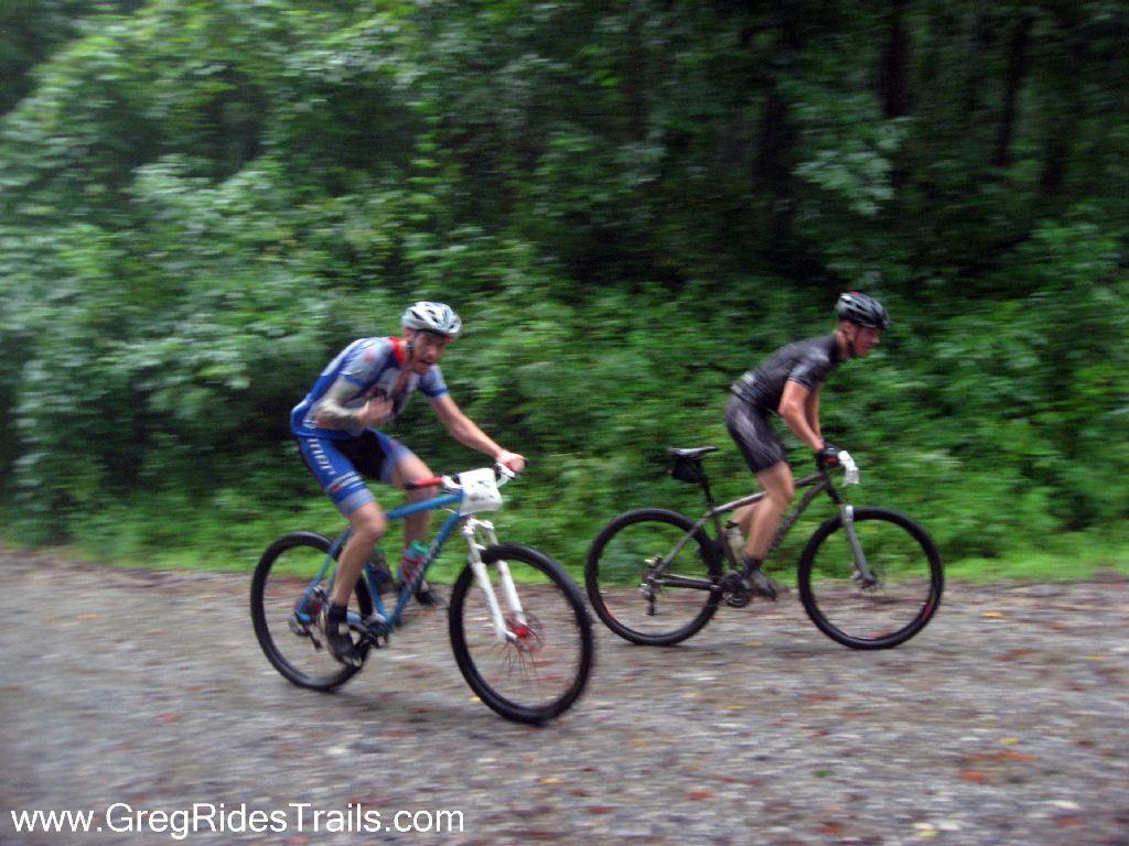 Two mountain bikers navigating a gravel trail during rainy weather, surrounded by dense greenery. The rider in the foreground is wearing a blue and red cycling outfit, while the rider in the background is dressed in black. Both cyclists appear focused as they tackle the muddy terrain. Winding Stairs Loop mountain bike trail.