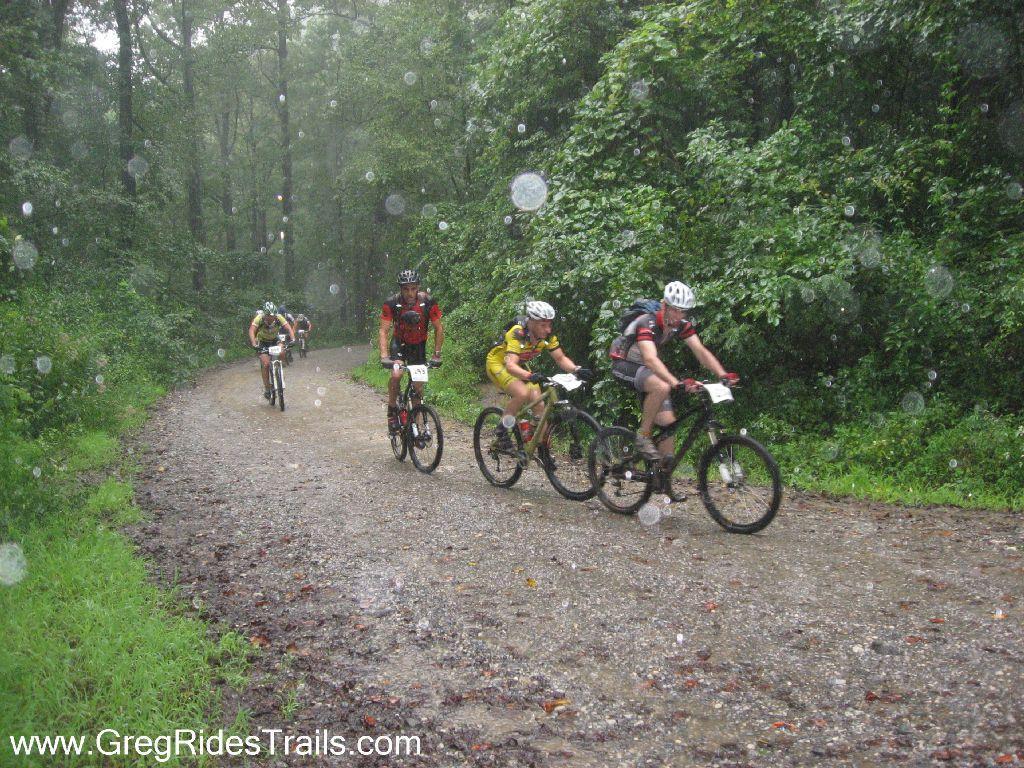A group of four mountain bikers riding on a gravel trail in rainy weather. The lush green forest surrounds the path, and rain droplets are visible in the air. The cyclists are wearing colorful jerseys and helmets, and some have race numbers attached to their clothing. Winding Stairs Loop mountain bike trail.