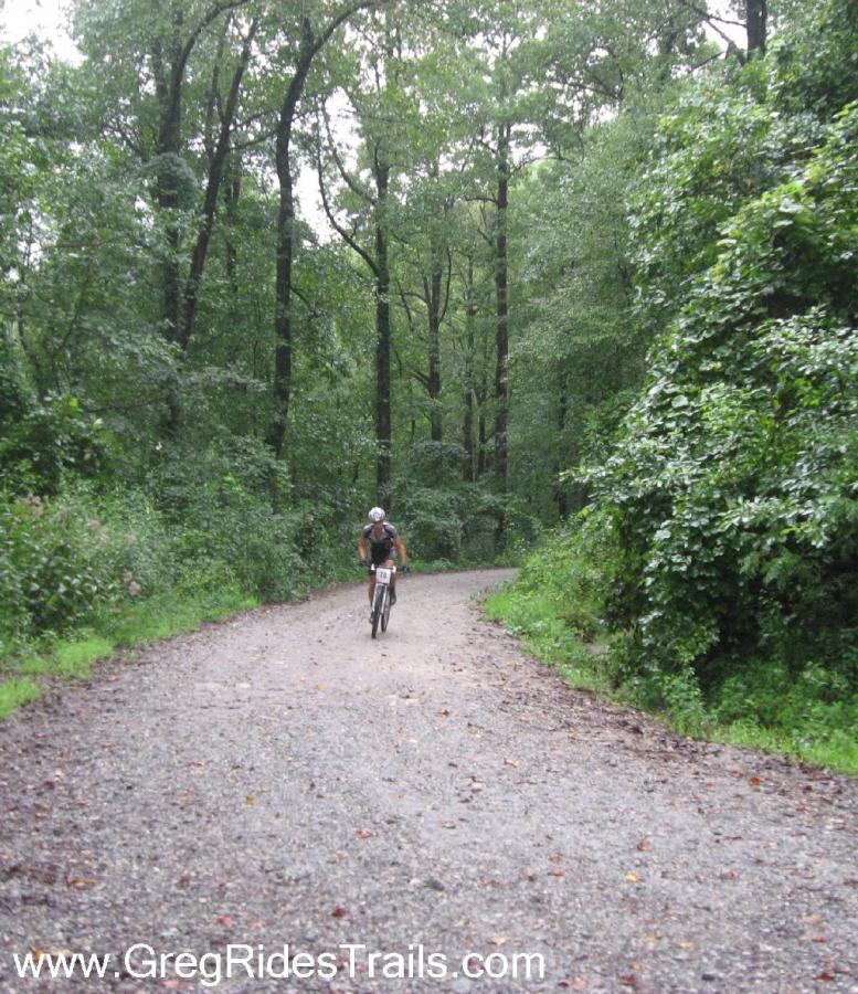 A cyclist riding on a gravel path surrounded by dense green trees in a forested area. The scene appears tranquil and lush, suggesting an outdoor recreational trail. Winding Stairs Loop mountain bike trail.