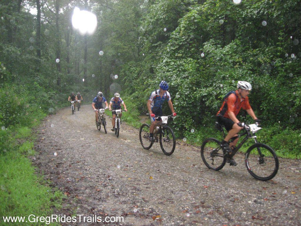 A group of mountain bikers riding on a gravel path in a dense, rain-soaked forest. The cyclists are equipped with helmets and jerseys, navigating through the wet conditions as raindrops are visible in the air. Lush greenery surrounds the trail, creating a vibrant, natural backdrop. Winding Stairs Loop mountain bike trail.