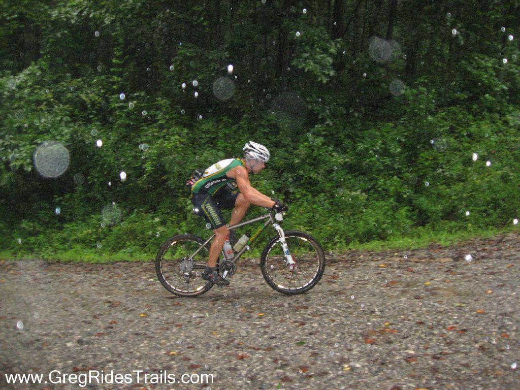 A cyclist riding a mountain bike on a gravel trail during a rainstorm, surrounded by lush green vegetation. Raindrops are visible in the air, creating a dynamic and adventurous atmosphere. Winding Stairs Loop mountain bike trail.