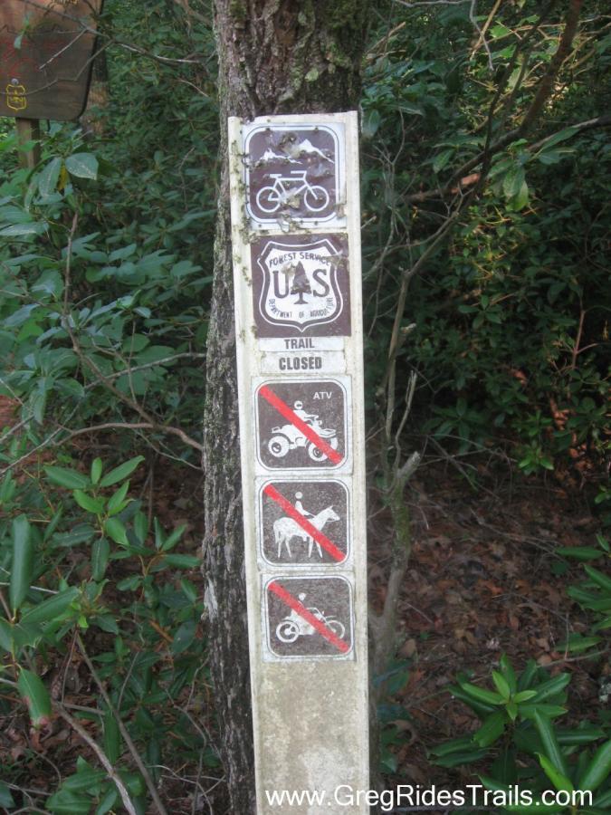 Signpost displaying various outdoor activity restrictions. The sign includes symbols for biking, indicating a closed trail, and prohibits the use of ATVs, horseback riding, and motorcycles. It is mounted on a tree surrounded by greenery. White Twister mountain bike trail.