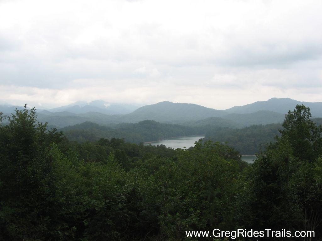 A scenic view of lush green mountains and a serene lake, under a cloudy sky. The landscape features layers of rolling hills fading into the distance, with trees and vegetation in the foreground. Tsali Recreation Area mountain bike trail.