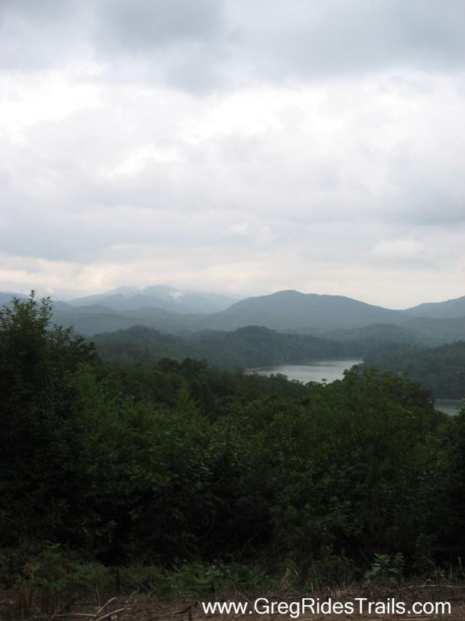 A scenic view of rolling mountains and a winding river, surrounded by lush greenery under a cloudy sky. The landscape conveys a sense of tranquility and natural beauty, ideal for outdoor enthusiasts. Tsali Recreation Area mountain bike trail.