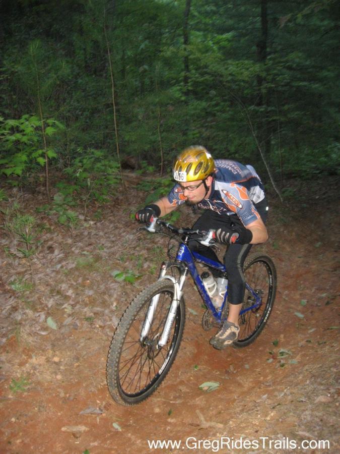 A cyclist in a yellow helmet and cycling gear is navigating a dirt trail through a wooded area. The rider leans forward, focused on the path ahead while riding a blue mountain bike. The surrounding environment features green foliage and a mix of dirt and gravel on the trail. Bull Mountain Cutoff / Fdr-83 / 223d mountain bike trail.