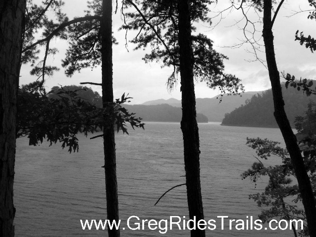 Black and white image of a serene lake surrounded by distant mountains, framed by the silhouettes of tall trees in the foreground. The water appears calm under a cloudy sky, creating a tranquil atmosphere. Tsali Recreation Area mountain bike trail.