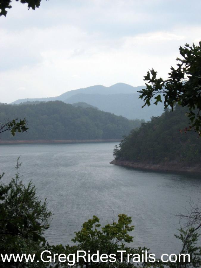 Scenic view of a lake surrounded by lush green hills and mountains under a cloudy sky. The water reflects the surrounding landscape, creating a tranquil atmosphere. Tsali Recreation Area mountain bike trail.