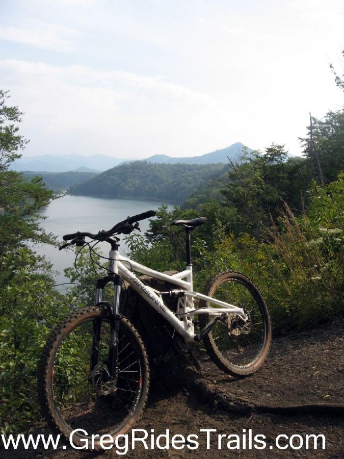 A mountain bike resting on the edge of a scenic overlook, surrounded by greenery, with a serene lake and mountains in the background under a cloudy sky. Tsali Recreation Area mountain bike trail.