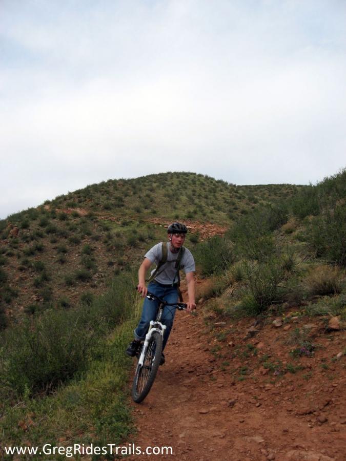 A mountain biker riding on a dirt trail through a green, hilly landscape under a cloudy sky. Devil's Backbone mountain bike trail.