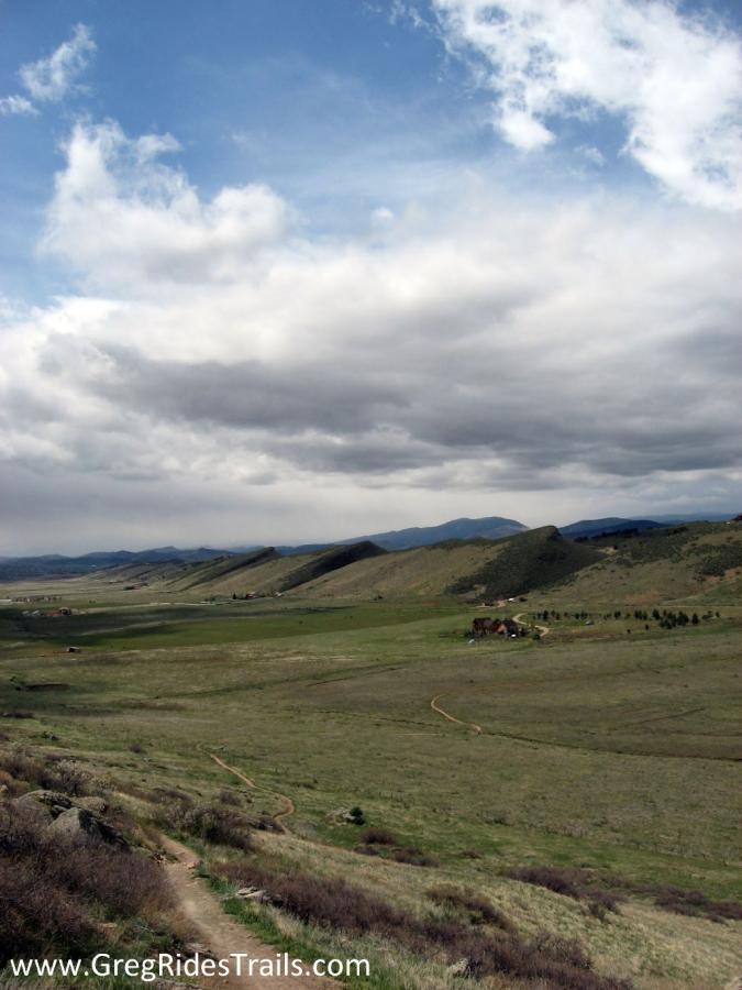 A panoramic view of rolling green hills under a cloudy sky, with a winding dirt trail leading through the foreground. In the distance, some small buildings and trees are visible, set against a backdrop of distant mountains. Coyote Ridge mountain bike trail.