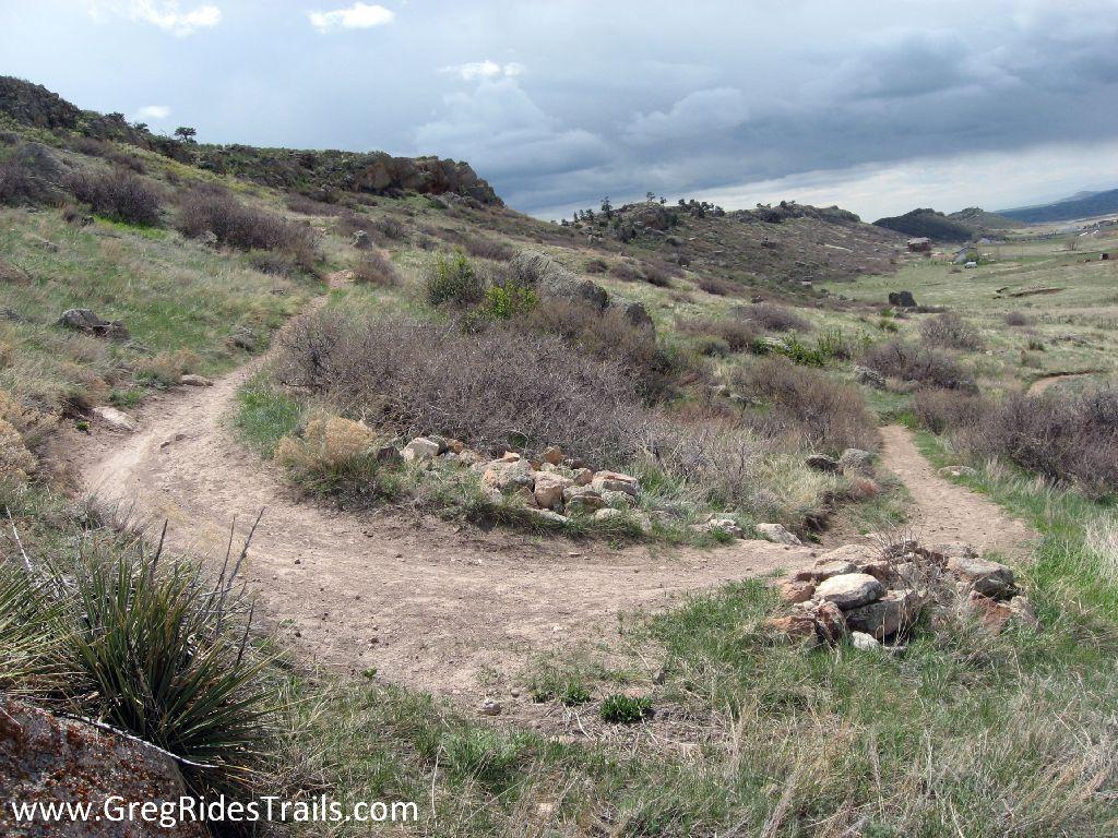 A scenic view of a winding dirt trail through grassy terrain, surrounded by shrubs and scattered rocks, under a cloudy sky with hints of blue. Coyote Ridge mountain bike trail.