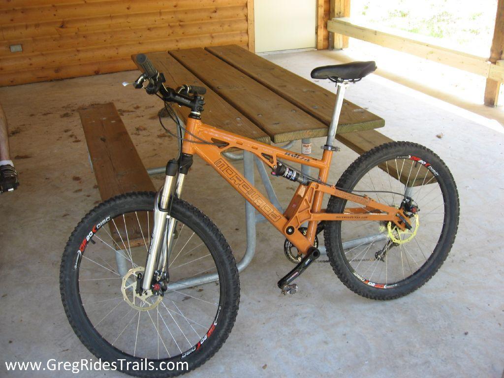 A mountain bike with an orange frame and thick tires rests beside a wooden picnic table in a rustic outdoor shelter. The bike features front suspension and a sleek design, showcasing its suitability for off-road trails. Levis Mounds mountain bike trail.