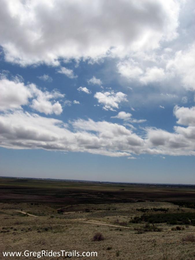 A wide view of an open landscape under a blue sky filled with fluffy white clouds. The foreground features rolling hills with sparse vegetation, while the horizon stretches far into the distance. A winding path is visible leading through the grassy terrain, evoking a sense of tranquility and natural beauty. Coyote Ridge mountain bike trail.