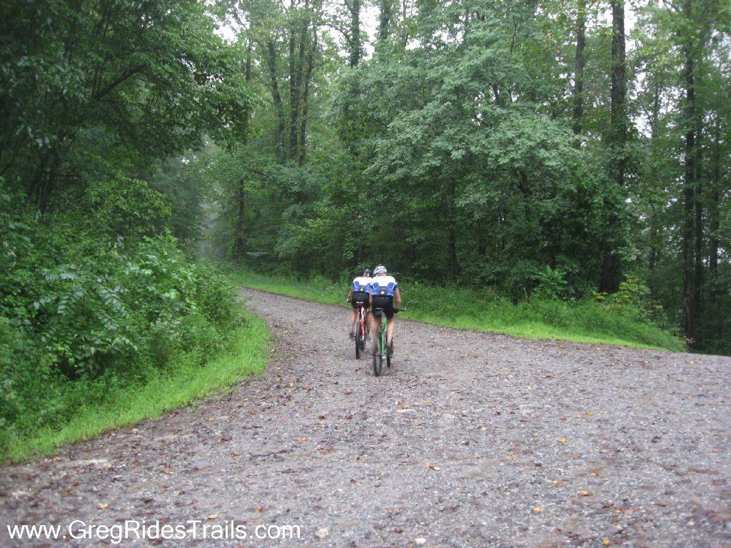 Two cyclists riding on a gravel trail surrounded by lush green trees on a rainy day. The trail curves ahead, leading into a misty, forested area. Winding Stairs Loop mountain bike trail.