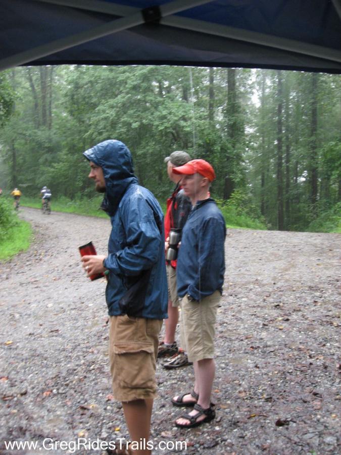 Three people standing under a canopy in a rainy outdoor setting, wearing rain gear and looking at the wet, gravel path ahead. Trees and greenery are visible in the background, indicating a forested area. One person is holding a red cup, while another has a water bottle. Their expressions suggest they are observing an activity occurring in the distance. Winding Stairs Loop mountain bike trail.