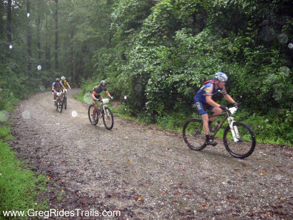 Mountain bikers racing on a gravel trail in a forest during rainy weather, surrounded by lush green foliage. Winding Stairs Loop mountain bike trail.