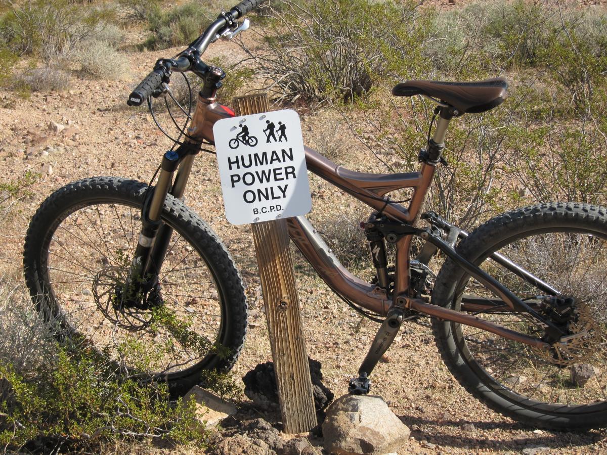 A mountain bike leaning against a wooden post with a sign that reads "HUMAN POWER ONLY," set in a natural outdoor environment with sparse vegetation and rocky terrain. Bootleg Canyon mountain bike trail.